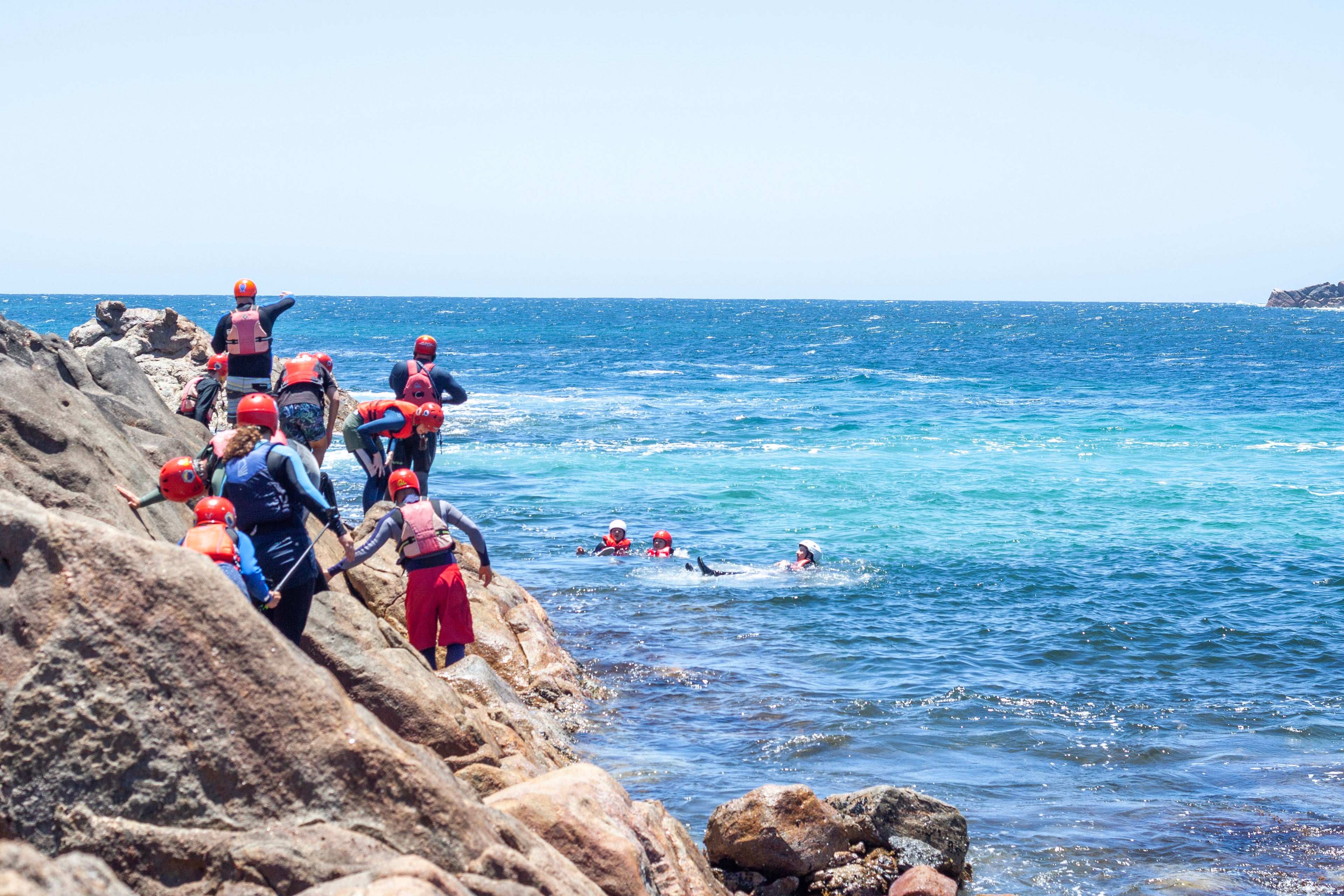 A group of people in helmets and life jackets on a rocky coastline, some getting into the sea